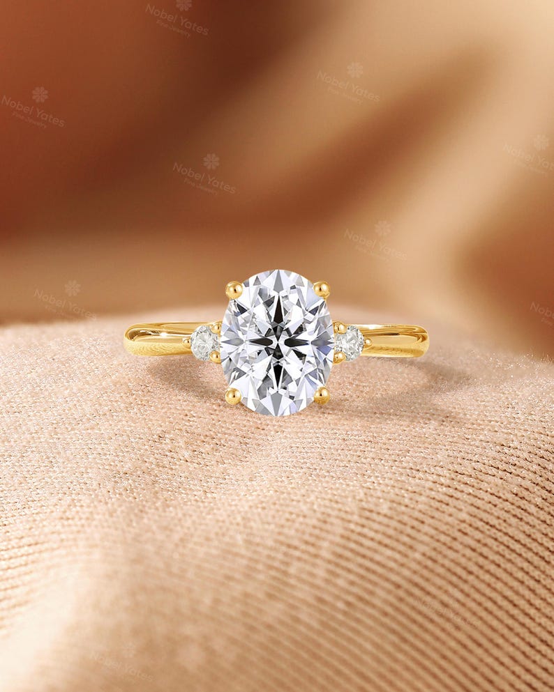 May include: A yellow gold ring featuring a large oval-cut center stone held by four prongs, flanked by a single smaller round-cut stone on each side. The band is smooth and polished, showcasing a classic three-stone design with clear, faceted stones.