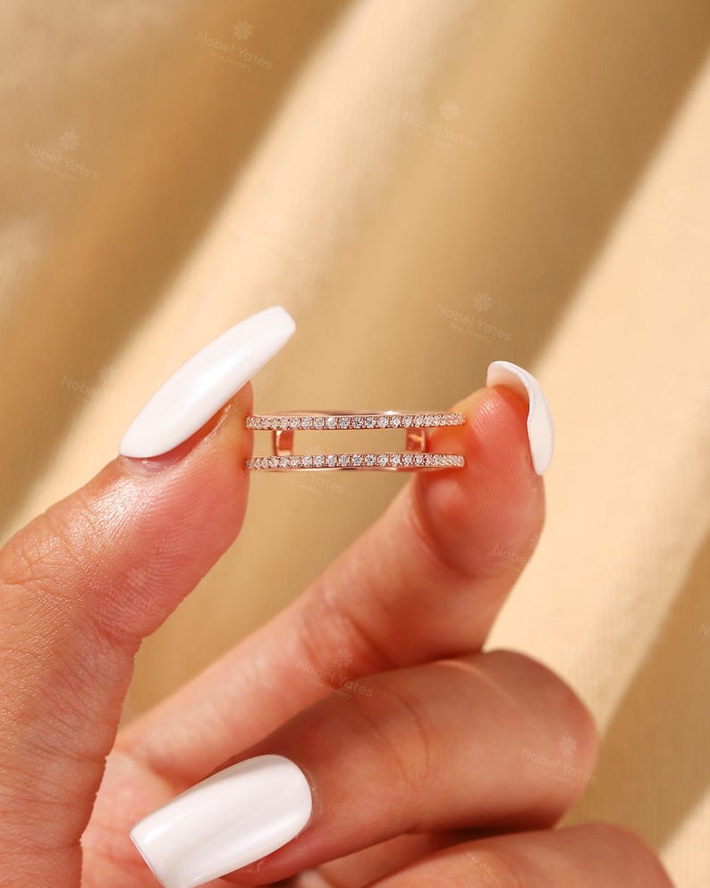 May include: A rose gold double band ring featuring two parallel rows of small, clear, pav&eacute;-set stones. The ring is constructed with two thin metal bars connecting the bands on each side, creating an open space between the two circular bands.