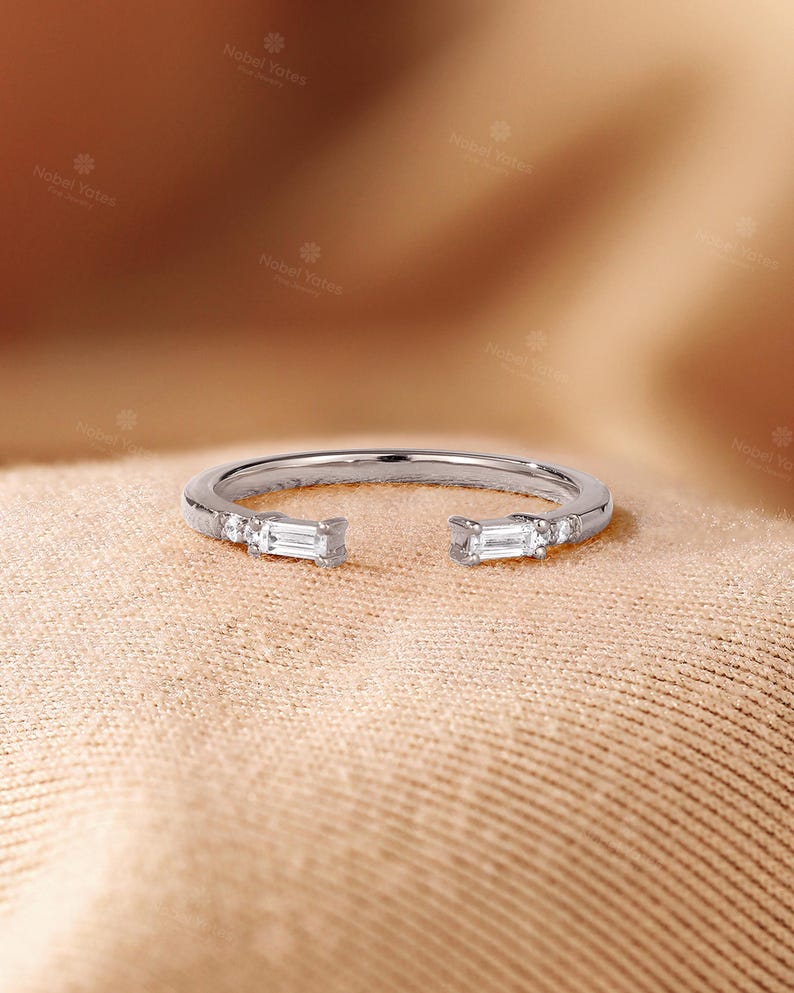 May include: A silver ring with an open design, featuring two rectangular-cut stones at the ends and smaller stones along the band. The ring is set against a neutral, textured background. The brand name "Nobel Yates" is visible.
