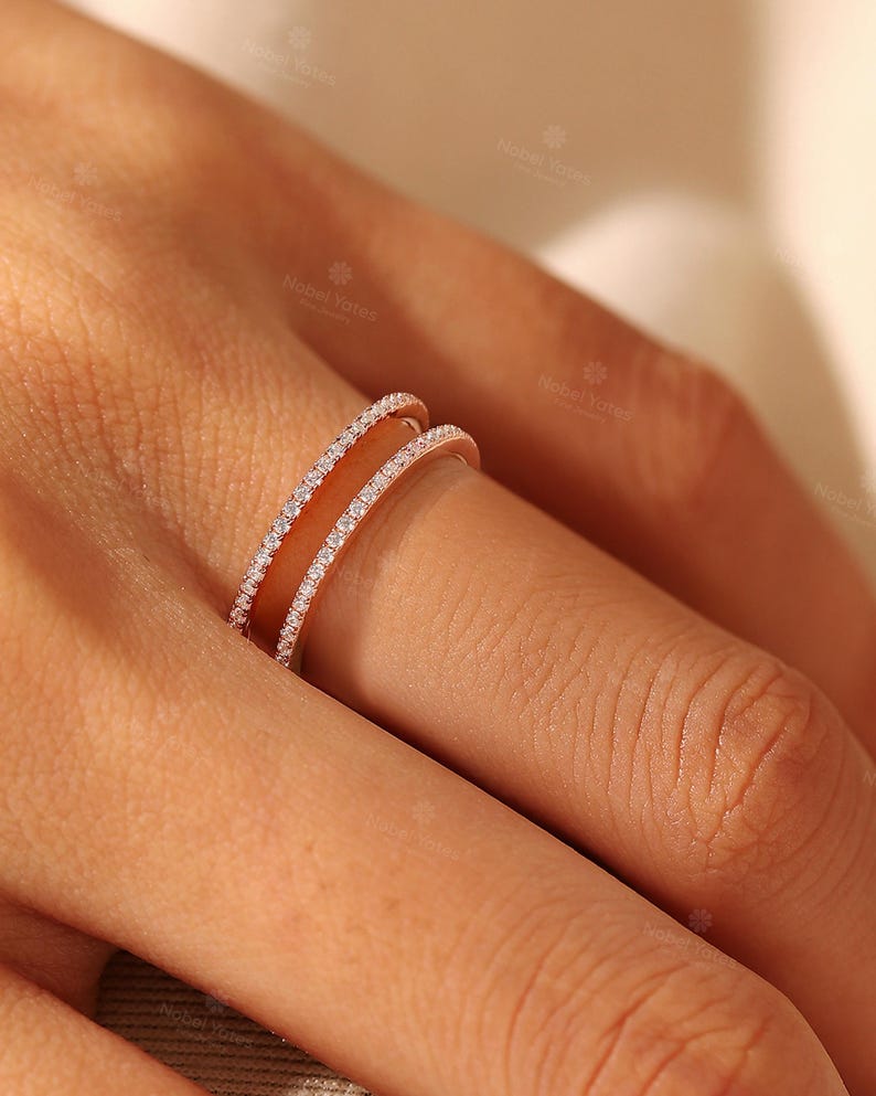 May include: A rose gold ring featuring two parallel bands connected by a bridge, with each band encrusted with a row of small, clear, round-cut stones. The design creates an open space between the two bands, showcasing a metallic finish.