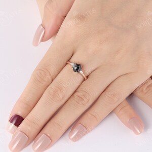 Pode incluir: Um anel de noivado de ouro rosa com uma pedra central de diamante preto e dois diamantes brancos menores de cada lado. O anel est&aacute; em um dedo com esmalte de unha rosa claro.