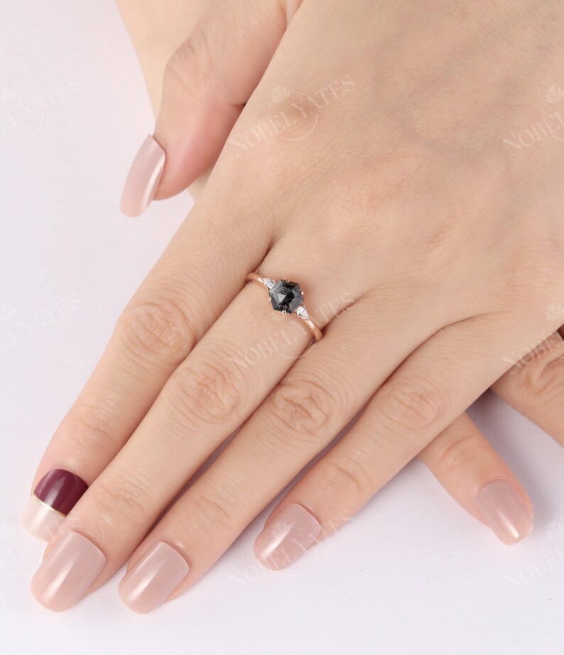 Pode incluir: Um anel de noivado de ouro rosa com uma pedra central de diamante preto e dois diamantes brancos menores de cada lado. O anel est&aacute; em um dedo com esmalte de unha rosa claro.