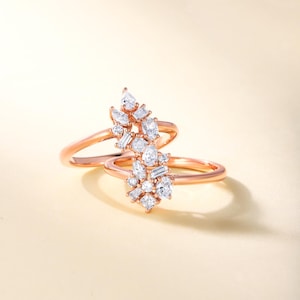 May include: Two rose gold rings, each featuring a cluster of white gemstones. The gemstones are a mix of round, pear, and baguette cuts, forming a sparkling floral pattern. The rings are displayed against a soft, neutral backdrop.