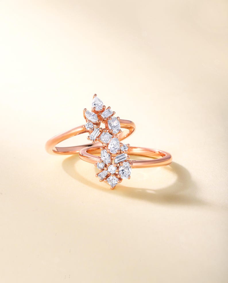 May include: Two rose gold rings, each featuring a cluster of white gemstones. The gemstones are a mix of round, pear, and baguette cuts, forming a sparkling floral pattern. The rings are displayed against a soft, neutral backdrop.