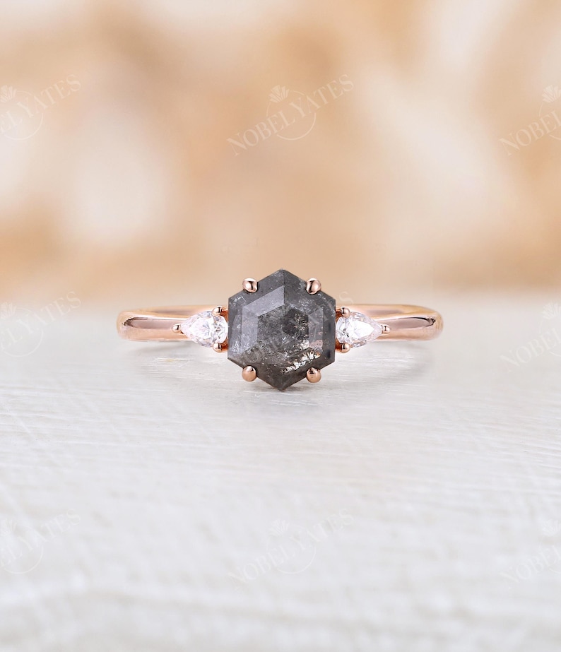 Pode incluir: Um anel de noivado de ouro rosa com um diamante hexagonal cinza e dois diamantes de acentua&ccedil;&atilde;o em forma de p&ecirc;ra.