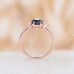 Pode incluir: Um anel de noivado em ouro rosa com uma pedra central de diamante preto e dois diamantes menores de cada lado.
