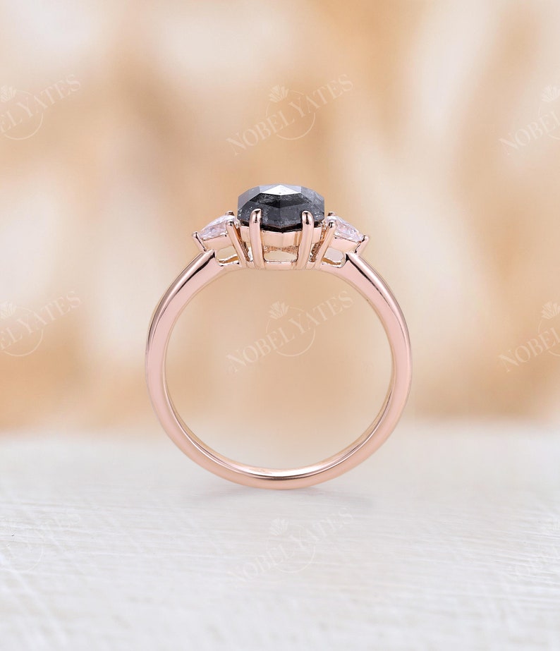 Pode incluir: Um anel de noivado em ouro rosa com uma pedra central de diamante preto e dois diamantes menores de cada lado.