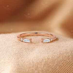 May include: Rose gold open ring with baguette and round cut diamonds. The ring features two baguette diamonds on each end, with smaller round diamonds set along the band. The ring is displayed on a beige fabric.