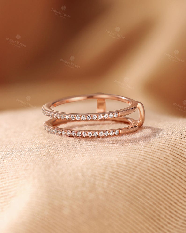 May include: A rose gold double band ring featuring two parallel bands connected by a small vertical bar. Both bands are encrusted with a row of small, clear, round-cut stones along the outer surface, creating a delicate and refined appearance.