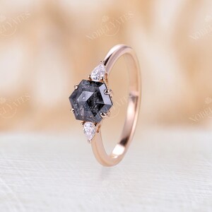 Pode incluir: Um anel de noivado de ouro rosa com um diamante preto hexagonal e dois diamantes menores em forma de p&ecirc;ra nas laterais.