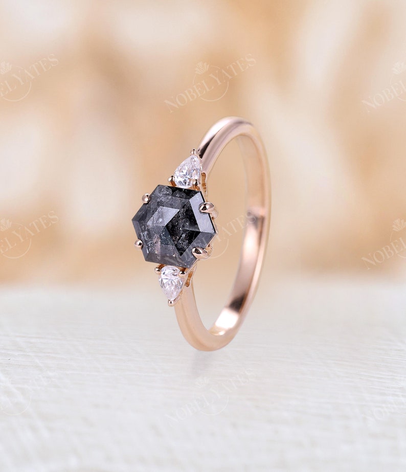 Pode incluir: Um anel de noivado de ouro rosa com um diamante preto hexagonal e dois diamantes menores em forma de p&ecirc;ra nas laterais.