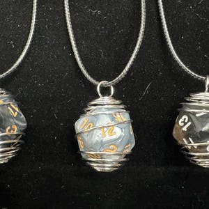 Puede incluir: Tres collares de jaula de alambre plateado con un colgante de dados blanco y negro. Cada dado tiene números dorados.