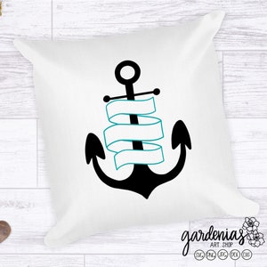 Puede incluir: Cojín cuadrado blanco con un diseño de ancla negra y una pancarta turquesa. El cojín está decorado con un tema náutico. El cojín está sobre una superficie de madera blanca con piñas y piedras decorativas.