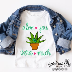 Könnte beinhalten: Weißes T-Shirt mit einer Illustration einer grünen Aloe-Vera-Pflanze und dem Text "aloe you vera much" in grüner Schrift mit rosa Herzen.