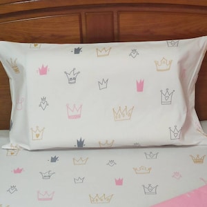 May include: A white and pink bedding set with a crown pattern. The pillowcase features a white background with gold, pink, and gray crowns. The fitted sheet is pink with white crowns.