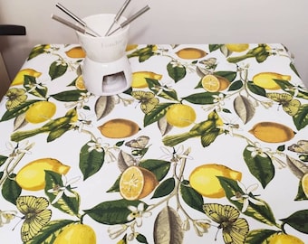 May include: A white ceramic fondue pot with stainless steel forks sits on a table covered with a white tablecloth. The tablecloth features a lemon and bird pattern with yellow lemons, green leaves, and yellow butterflies.