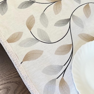 Puede incluir: Un camino de mesa blanco con un delicado ribete de encaje presenta un patrón repetitivo de hojas marrones y grises. El camino de mesa está colocado sobre una mesa de madera oscura con un plato blanco.