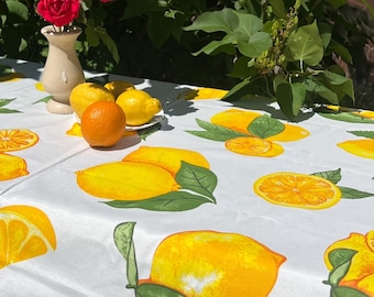 May include: A white tablecloth with a vibrant lemon pattern, featuring whole and sliced lemons with green leaves. A vase of red roses and a plate of lemons and an orange sit on the table. The scene is outdoors.