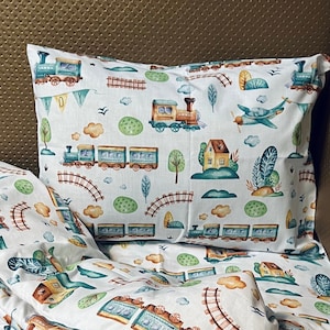 May include: White bedding with a train and airplane pattern. The bedding features a green, blue, and brown train with a yellow and brown house in the background. The bedding also features a blue airplane with white wings.