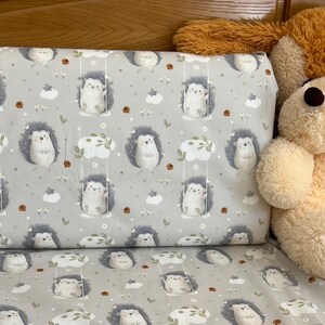 May include: A light gray baby bedding set featuring a repeating pattern of cartoon hedgehogs on swings, clouds, and flowers. The set includes a pillow and a sheet, with a stuffed animal visible in the background.