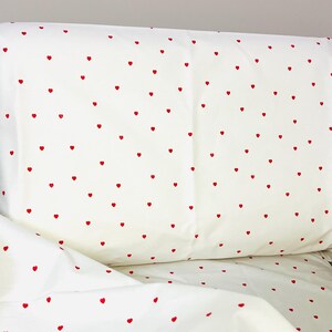 Peut inclure: Ensemble de literie blanc avec un motif répété de petits cœurs rouges. L'ensemble comprend une taie d'oreiller et un drap, tous deux affichant le motif de cœur. Le tissu semble doux et lisse, adapté à une chambre.