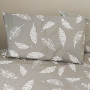 May include: A grey bedspread with a white feather pattern. Two matching pillowcases with the same feather pattern are on top of the bedspread.