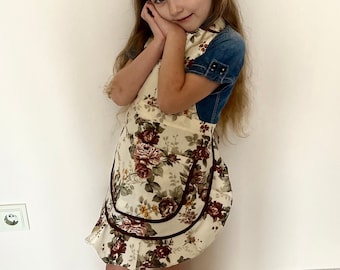 May include: A young girl wearing a floral print apron with a brown trim and a denim top. She is standing in front of a white wall with a light switch.