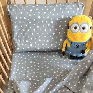 May include: A gray crib sheet with a white star pattern. A yellow plush minion toy is sitting on the sheet.