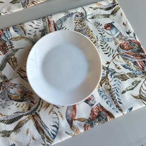 Puede incluir: Un juego de cuatro platos de cerámica blanca con fondo crema y un estampado colorido de pájaros y hojas. Los platos están sobre una mesa con un mantel a juego.