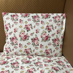 Könnte beinhalten: Weißes Kissen und Bettwäsche-Set mit einem sich wiederholenden Muster aus rosa und roten Rosen und gelben Akzenten. Das florale Design ist vor einem weißen Hintergrund angeordnet. Das Bettwäsche-Set ist wahrscheinlich aus einem weichen Stoff.