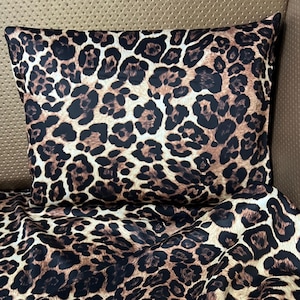 Może przedstawiać: Zestaw poduszek i pościeli z nadrukiem w cętki. Wzór zawiera brązowe i czarne plamy na beżowym tle. Tkanina wydaje się miękka i luksusowa, odpowiednia do dekoracji sypialni.