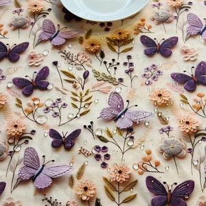 Può includere: Una tovaglia bianca con un motivo floreale con farfalle viola e piccoli fiori rosa e marroni. La tovaglia ha un piatto bianco al centro.