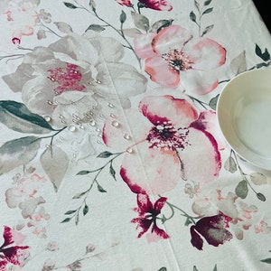 May include: White tablecloth with a floral pattern featuring pink and burgundy flowers with green leaves. Water droplets are visible on the surface of the tablecloth.