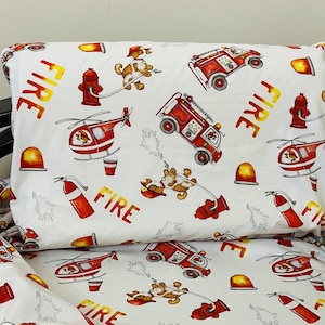 May include: White bedding with a red, yellow and black fire engine, helicopter and dog pattern. The bedding has the word "FIRE" repeated throughout the pattern.
