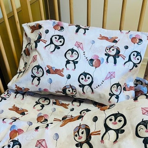 May include: White baby bedding set with a playful penguin theme. The set includes a pillowcase and a fitted sheet, both adorned with cartoon penguins, airplanes, kites, and balloons in shades of brown, pink, and blue.