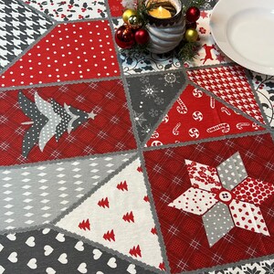 Peut inclure: Une nappe de table en patchwork rouge et gris sur le thème de Noël. La nappe présente différents motifs, notamment des flocons de neige, des cannes en sucre, du houx et des arbres. Une assiette blanche et un bougeoir avec des ornements rouges et dorés sont sur la nappe.