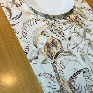 Puede incluir: Un camino de mesa blanco con un patrón de pájaros y hojas tropicales marrones y verdes. Un plato blanco está colocado sobre el camino de mesa.