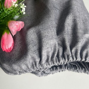 Op de afbeelding: Een grijze stoffen tas met een geplooide elastische rand. De tas is versierd met een boeket roze bloemen en groene bladeren.