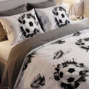 Op de afbeelding: Een wit dekbedovertrek en twee bijpassende kussenslopen met een zwart-wit voetbal- en voetbalschoenpatroon met inktspatontwerpen. De beddengoedset is opgemaakt op een bed met grijze kussens en een grijs hoeslaken in een slaapkamer.