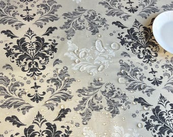 Puede incluir: Un mantel beige con un patrón de damasco repetido en blanco y negro. Se ven gotas de agua en la superficie. Un plato blanco está en la esquina superior derecha. El mantel es probablemente resistente al agua.