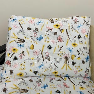 Könnte beinhalten: Weißes Kissenbezug- und Bettlaken-Set mit einem farbenfrohen Blumen- und Insektenmuster. Das Design umfasst gelbe Sonnenblumen, rosa Blüten, blaue Libellen, Schmetterlinge und Bienen. Das Bettwäsche-Set ist aus einem weichen, leichten Stoff gefertigt.