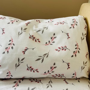 May include: White pillowcase and bedding set with a repeating pattern of gray leaves and pink-red berries. The pillow is rectangular and rests on a matching sheet. The background is a neutral beige.