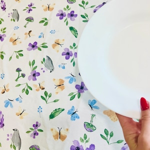 May include: White tablecloth with a floral pattern featuring butterflies, birds, and mushrooms. Two white plates are on the tablecloth, one is being held by a hand.
