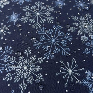May include: A dark navy blue fabric featuring a repeating pattern of white and light blue snowflakes of varying sizes. The surface of the material is covered in small, clear water droplets that sit on top of the printed winter design.