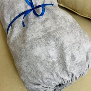 Könnte beinhalten: Ein grauer Stoffartikel, möglicherweise ein Kissenbezug oder eine Hülle, der mit einem blauen Band gebunden ist. Der Stoff hat ein gesprenkeltes Muster und der Artikel ist an einem Ende gerafft. Der Hintergrund ist eine hellbeige Oberfläche.