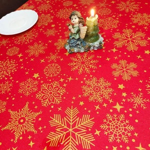 May include: Red tablecloth with a gold snowflake pattern and small gold stars. A small angel figurine with a lit candle is in the center of the tablecloth.