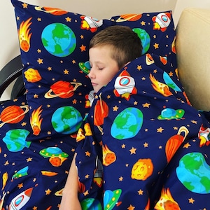 Può includere: Un bambino dorme sotto un copripiumino blu e arancione a tema spaziale con pianeti, stelle e razzi. Il copripiumino è realizzato in tessuto morbido e confortevole.