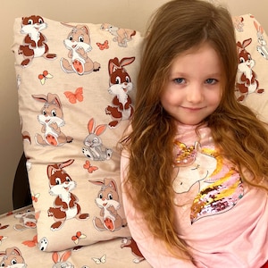 May include: A young girl with long brown hair is sitting on a couch with a pillow featuring a pattern of brown bunnies and butterflies. She is wearing a pink shirt with a sequined unicorn design.