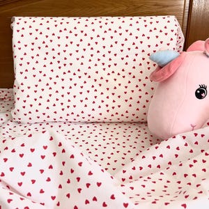 Peut inclure: Ensemble de literie blanc orné de petits cœurs rouges. L'ensemble comprend une taie d'oreiller et un drap, avec un jouet en peluche licorne rose avec une corne bleue et une crinière rose. La literie est sur un cadre de lit en bois.