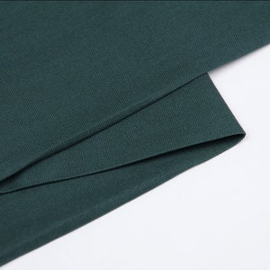 May include: Close-up of a dark green fabric with a textured weave. The fabric is folded in thirds, showing the smooth, even texture.